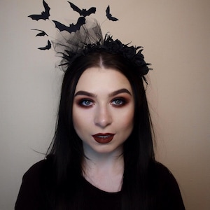 May include: A black Halloween headpiece featuring a floral crown, black tulle, and several black bat cutouts. The bats are in various sizes and positioned to appear as if they are flying. The person in the image is wearing dark makeup and a black top.