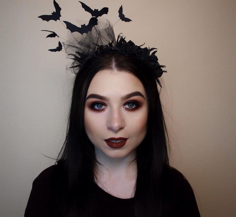 May include: A black Halloween headpiece featuring a floral crown, black tulle, and several black bat cutouts. The bats are in various sizes and positioned to appear as if they are flying. The person in the image is wearing dark makeup and a black top.