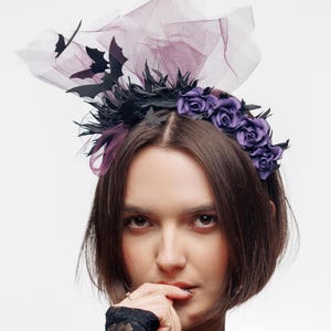 May include: A headband featuring a cluster of deep purple fabric roses, black bat-shaped cutouts, and a layer of sheer, dark purple tulle netting that extends upward. The accessory is designed to be worn on the head as a decorative hair piece.