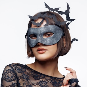 May include: A grey textured mask with a bat design, complemented by a bat-themed headpiece. The mask covers the eyes and nose. The person is wearing a black lace top. Ideal for Halloween or fancy dress events.