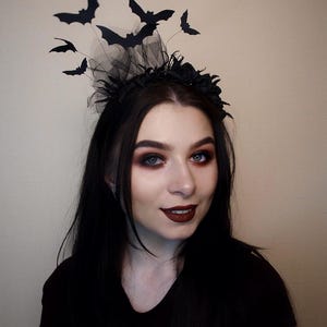 May include: A black Halloween headband featuring a black tulle base, black floral accents, and several black bat cutouts. The bats are attached to thin wires, giving the appearance of flight. The headband is a festive accessory for Halloween.
