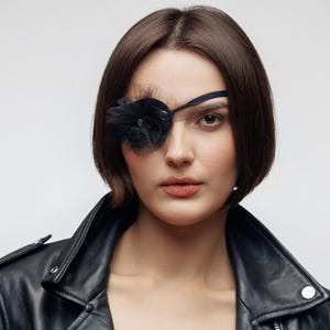 May include: A woman with short brown hair wearing a black leather jacket and a black eye patch. The eye patch is adorned with a black flower and tulle. The background is a plain white.
