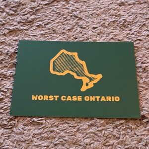 May include: A green rectangular card with the words "WORST CASE ONTARIO" in gold lettering. The card features a gold outline of the province of Ontario.