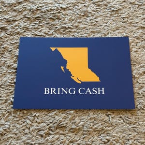 May include: A navy blue rectangular card with a gold outline of British Columbia, Canada. The text "BRING CASH" is printed in white below the map outline. The card is resting on a textured, light brown surface.