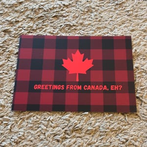 May include: A rectangular card with a red and black plaid pattern. A red maple leaf is centered on the card. The text "GREETINGS FROM CANADA, EH?" is printed in red below the maple leaf.