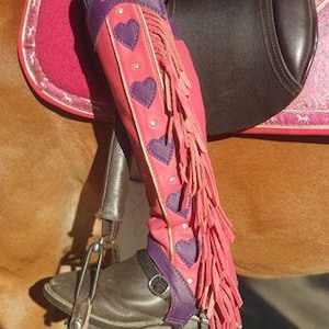 May include: Pink and purple riding boots with heart accents and fringe detail. The boots have the name "Sabrina" embroidered on the top. The boots are worn with a black stirrup and a brown saddle.