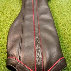 May include: Black leather item with red trim and stitching, possibly a boot or a protective sleeve. The item has a curved shape and is set against a green, grassy background. The red trim adds a striking contrast to the black leather.