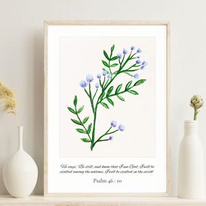 以下が含まれることがあります： 緑の葉と小さな青い花を持つ、開花植物の植物図が描かれた、額入りの水彩画プリント。アートワークには、詩篇46:10からの引用が含まれています。
