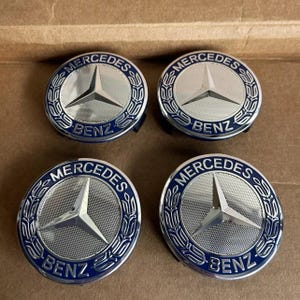 以下が含まれることがあります： 4つのシルバーとブルーのメルセデスベンツホイールセンターキャップ。各キャップには、メルセデスベンツのロゴ、円形エンブレム内のスリーポインテッドスター、および「BENZ」の文字が表示されています。キャップは正方形に配置されています。
