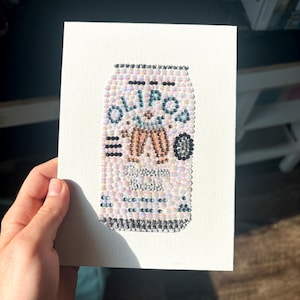 May include: A canvas artwork featuring a beaded design of an Olipop soda can. The can is primarily light pink with the brand name in blue and other details in brown and black. The artwork is held by a hand.