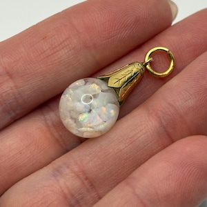 May include: A gold-colored pendant with a round, clear orb filled with white, iridescent fragments. The pendant has a floral-shaped top and a small loop for a chain. The orb appears to be made of glass or crystal.