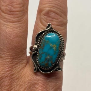 May include: A silver ring featuring an oval-shaped turquoise stone with brown matrix veining. The stone is set within a twisted rope border and accented by decorative silver scrollwork on the sides of the band, creating a detailed metal design.