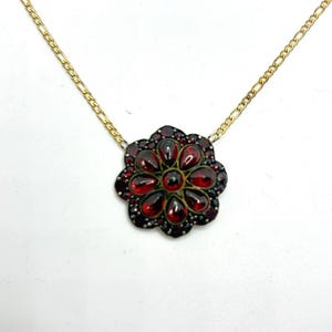 May include: A gold-toned chain necklace featuring a central floral-shaped pendant. The pendant is decorated with multiple deep red, teardrop-shaped stones arranged in a circular pattern, surrounded by smaller dark red stones set in a dark metal frame.