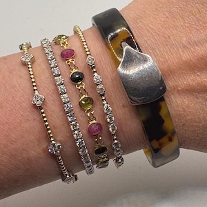 May include: An assortment of bracelets on a wrist. The bracelets include a gold chain with floral accents, a tennis bracelet, a gold chain with gemstones, and a silver chain with diamonds. A wide, dark brown and yellow bangle with a silver clasp is also present.