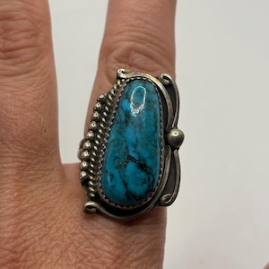 Op de afbeelding: Een ring van sterling zilver met een grote, langwerpige, druppelvormige turkoois steen met donkere matrixaders. De zetting bevat een rand van kralendraad aan de ene kant en gladde, gebogen zilveren accenten aan de andere kant van de steen.