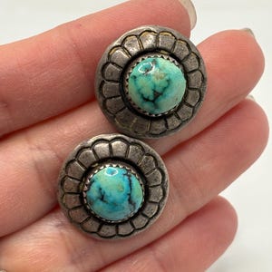 Op de afbeelding: Een paar zilverkleurige oorstekers met turkooizen stenen. Elke oorbel heeft een ronde, geschulpte rand rond een levendige, blauwgroene steen. De oorbellen worden op een hand getoond.