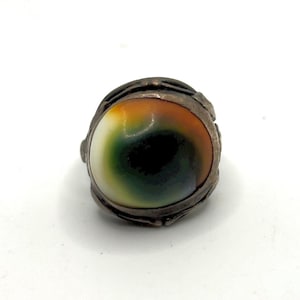 May include: A ring featuring a smooth, rounded cabochon stone with a gradient of white, green, and orange colors. The stone is set within a dark, textured metal band that wraps around the edges, creating a secure and detailed frame for the center.