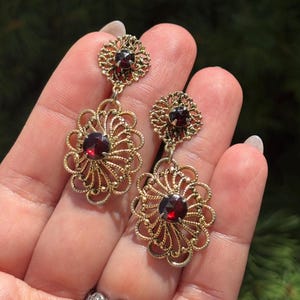 May include: A pair of gold-tone drop earrings featuring a circular filigree top and a larger, oval-shaped filigree base, each set with a round, deep red faceted gemstone in the center. The intricate metalwork creates a delicate, openwork pattern.