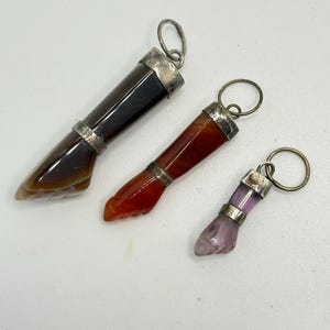 Könnte beinhalten: Drei Fuß-Anhänger aus Edelstein. Die Anhänger sind braun, rot und lila, mit silberfarbenen Metallakzenten und einer Schlaufe für eine Kette. Die Edelsteine haben eine polierte Oberfläche.