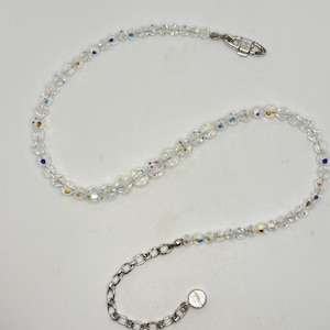 Puede incluir: Un collar de cristal transparente con acentos iridiscentes. El collar presenta pequeñas cuentas facetadas ensartadas, con un cierre plateado y una cadena ajustable. Las cuentas capturan la luz, mostrando un arcoíris de colores.