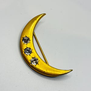 Peut inclure: Broche en forme de croissant de lune doré, ornée de trois pierres précieuses claires et étincelantes. La broche est munie d'une épingle au dos. Un accessoire de bijouterie sur le thème céleste.
