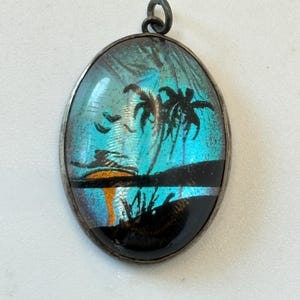 May include: Oval pendant with a silver-coloured frame. The pendant features a vibrant blue and black scene with palm trees, birds, and a sunset. The design is reminiscent of a tropical landscape.