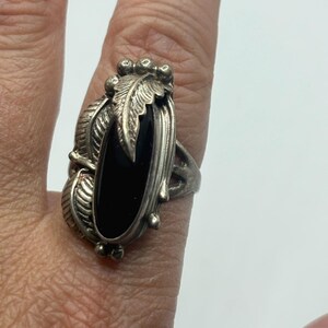 May include: A silver ring featuring a central vertical oval black stone. The metal setting is decorated with detailed leaf motifs on the sides and small spherical accents at the top and bottom, creating a textured, ornate appearance around the stone.