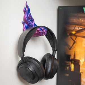 Könnte beinhalten: Schwarzer Over-Ear-Kopfhörer, der an einem Wand-Kopfhörerhalter hängt. Der Halter ist ein Flammen-Design in Lila und Blau. Die Kopfhörer haben große Ohrmuscheln.