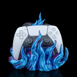 Könnte beinhalten: Ein weißer Gamecontroller ruht auf einem blauen, flammenartigen Sockel. Der Controller hat schwarze Akzente und Knopffdetails. Der Sockel hat ein wirbelndes Design mit roten Akzenten. Der Hintergrund ist schwarz.