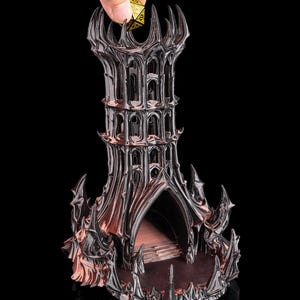 Könnte beinhalten: Ein detaillierter, dunkel gefärbter Würfelturm mit gotischem Architekturdesign. Der Turm hat eine spitze Spitze und gewölbte Öffnungen. Eine Hand hält einen goldfarbenen, vielseitigen Würfel über dem Turm.