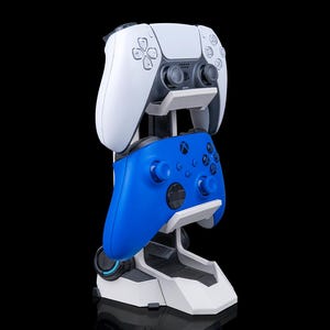 Könnte beinhalten: Ein weiß-blauer Gaming-Controller-Ständer, der einen weißen PlayStation 5-Controller und einen blauen Xbox-Controller hält. Der Ständer ist weiß und grau mit futuristischem Design, ideal für Gaming-Zubehör.