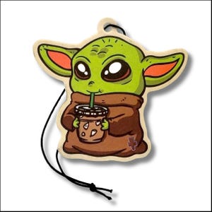May include: A green cartoon character with large eyes and pointed ears, holding a beverage with a straw. The character is wearing a brown robe and is an air freshener with a black string for hanging.