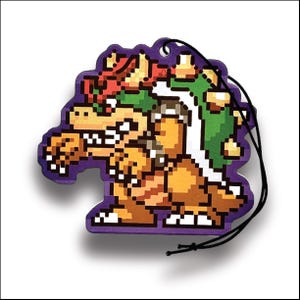 May include: A pixelated Bowser air freshener with a purple border and a black string for hanging. The design features Bowser's orange, green, and white color scheme, with a pixelated aesthetic reminiscent of classic video games.