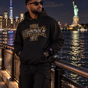 May include: A black hooded sweatshirt featuring the text THE BRONX NY in gold-outlined block letters with a cityscape graphic inside the lettering and a small gold crown icon above, plus a small Champion logo patch on the left sleeve cuff.