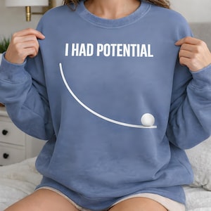 Pode incluir: Camisola azul com o texto branco "I HAD POTENTIAL". Abaixo do texto, há uma linha curva branca e um gráfico de bola branca. A camisola tem gola redonda e mangas compridas.