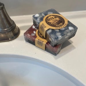 May include: Two rectangular bars of soap, one red and one dark gray, are stacked on a white surface. The gray soap has white speckles. Each bar is wrapped in clear plastic and has a brown paper label that reads "BITE ME" and "SOAP".
