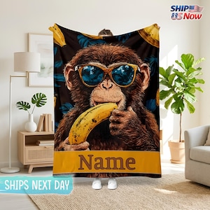 Könnte beinhalten: Eine braune Decke mit einem Affen, der eine Sonnenbrille trägt und eine Banane hält. Der Affe ist vor einem dunklen Hintergrund mit blauen floralen Akzenten und gelben Bananen platziert. Ein gelbes Banner am unteren Rand trägt das Wort "Name".