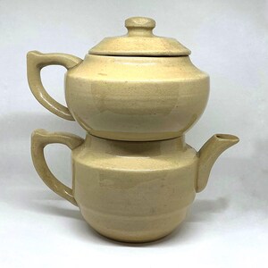 Könnte beinhalten: Ein zweiteiliges, hellbeige Keramik-Teekannen-Set. Die obere Kanne hat einen Deckel mit einem kleinen Knopf, und die untere Kanne hat einen Ausguss und einen Griff. Das Set ist gestapelt.
