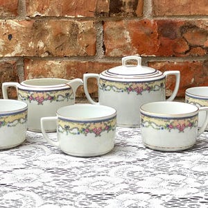 May include: A white porcelain tea set with floral and ribbon detailing. The set includes a lidded sugar bowl, a creamer, and four teacups. Each piece has a gold rim and a decorative band of pink, yellow, and blue floral elements.