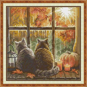 May include: Cross-stitch artwork depicting two cats gazing out a window at an autumn scene. The window reveals a colorful fall landscape with falling rain. A pumpkin and lit candles sit on the windowsill, with a lantern to the left.