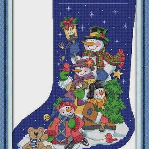 Pode incluir: Uma meia de Natal bordada em ponto cruz, emoldurada, com uma família de bonecos de neve, um ursinho de pelúcia e um passarinho. O fundo é azul escuro com flocos de neve brancos. Os bonecos de neve usam chapéus, cachecóis e casacos.