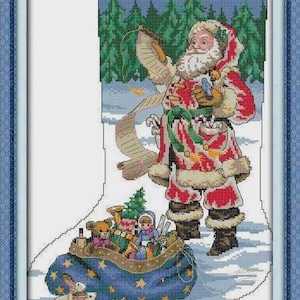 May include: Cross-stitch depiction of Santa Claus in a red and white suit, holding a scroll. A blue sack filled with toys, including teddy bears and dolls, sits at his feet. Two rabbits are in front of the sack. The background shows a snowy landscape and evergreen trees.