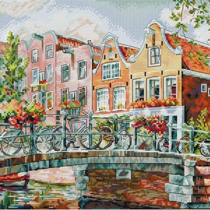 Peut inclure: Une broderie représentant une scène de canal européen. Des bâtiments colorés bordent le canal, avec des vélos garés sur un pont. Des fleurs rouges, oranges et jaunes ornent les balcons et le pont. Un petit bateau rouge est dans l'eau.