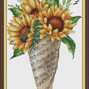 Puede incluir: Arte de punto de cruz de girasoles en un soporte en forma de cono hecho de partituras. Los girasoles tienen pétalos amarillos y centros marrones, con hojas verdes. La obra de arte está enmarcada en un marco marrón y dorado.