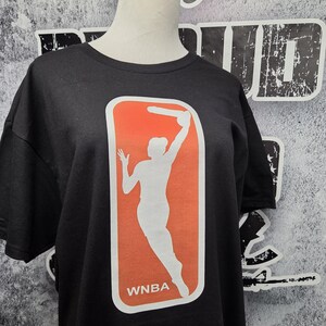 May include: Black t-shirt featuring the WNBA logo. The logo is a white silhouette of a basketball player in action, set against an orange rectangle with a white border. The letters "WNBA" are printed in white below the figure.