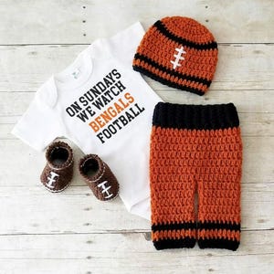 Può includere: Un body bianco a maniche corte con il testo nero e arancione ON SUNDAYS WE WATCH BENGALS FOOTBALL, abbinato a un berretto all'uncinetto arancione e nero, pantaloni coordinati arancione e nero e due scarpine all'uncinetto marroni con lacci bianchi.