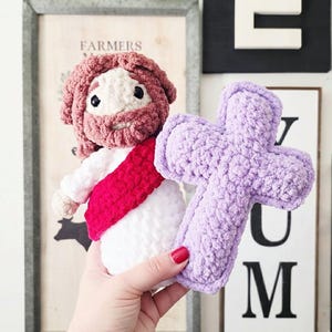 Puede incluir: Una mu&ntilde;eca de ganchillo hecha a mano que representa una figura con cabello y barba casta&ntilde;os, vestida con una t&uacute;nica blanca con una faja roja, sosteniendo una cruz de felpa de color morado claro. La mu&ntilde;eca est&aacute; hecha de hilo suave y texturizado.