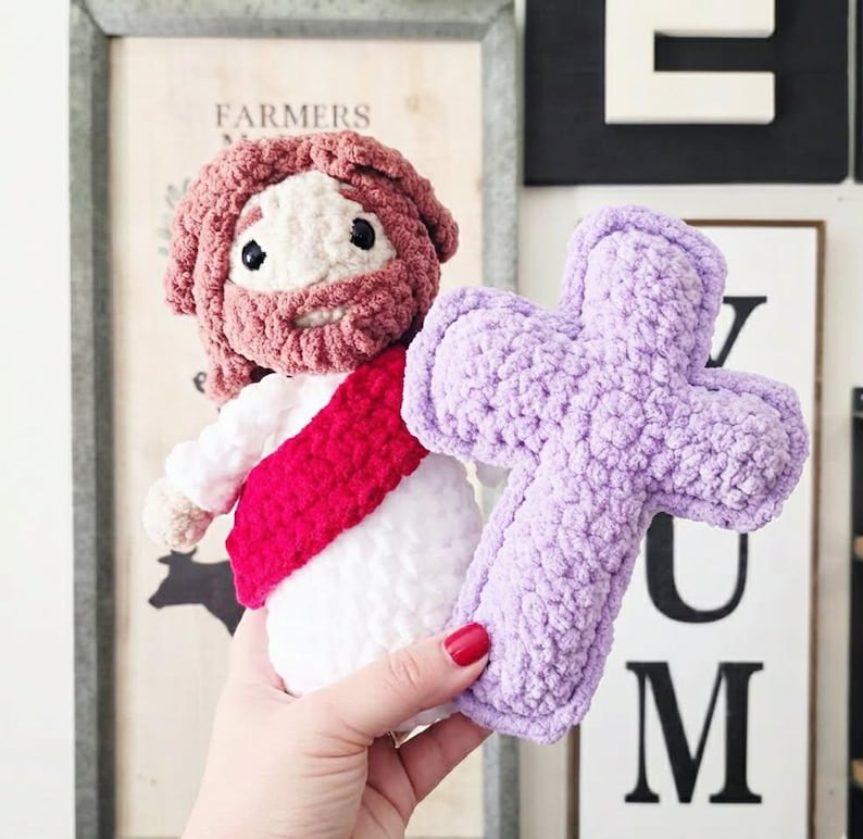 Puede incluir: Una mu&ntilde;eca de ganchillo hecha a mano que representa una figura con cabello y barba casta&ntilde;os, vestida con una t&uacute;nica blanca con una faja roja, sosteniendo una cruz de felpa de color morado claro. La mu&ntilde;eca est&aacute; hecha de hilo suave y texturizado.