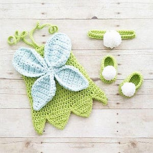May include: A crochet set consisting of a light green garment with a large, light blue, five-petaled flower applique and curly green tendrils, a matching light green headband with a white pom-pom, and two light green booties with white pom-poms.