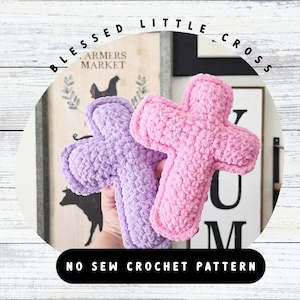 Puede incluir: Dos art&iacute;culos de felpa en forma de cruz hechos de hilo suave y texturizado en color morado claro y rosa brillante. La imagen incluye el texto BLESSED LITTLE CROSS y NO SEW CROCHET PATTERN. Estos art&iacute;culos presentan una construcci&oacute;n gruesa.